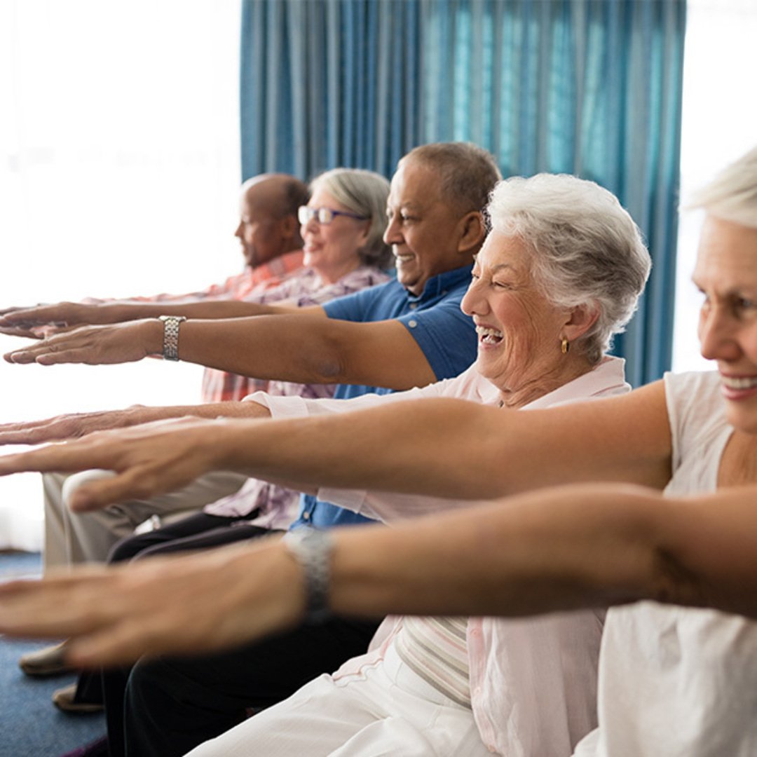 group of smiling people practising chair yoga with their arms stretched out
